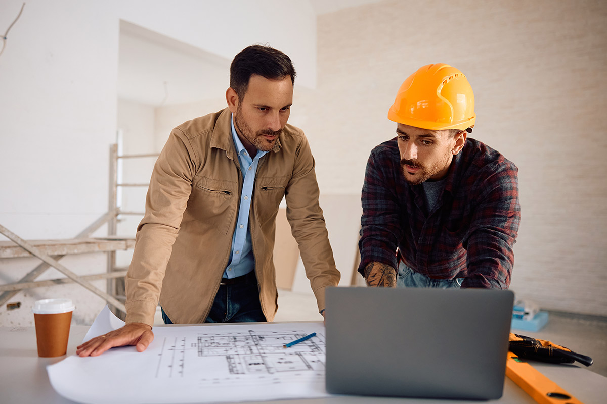 Two men wearing hard hats analyze data on a laptop, focusing on the responsibilities of an owner's representative.