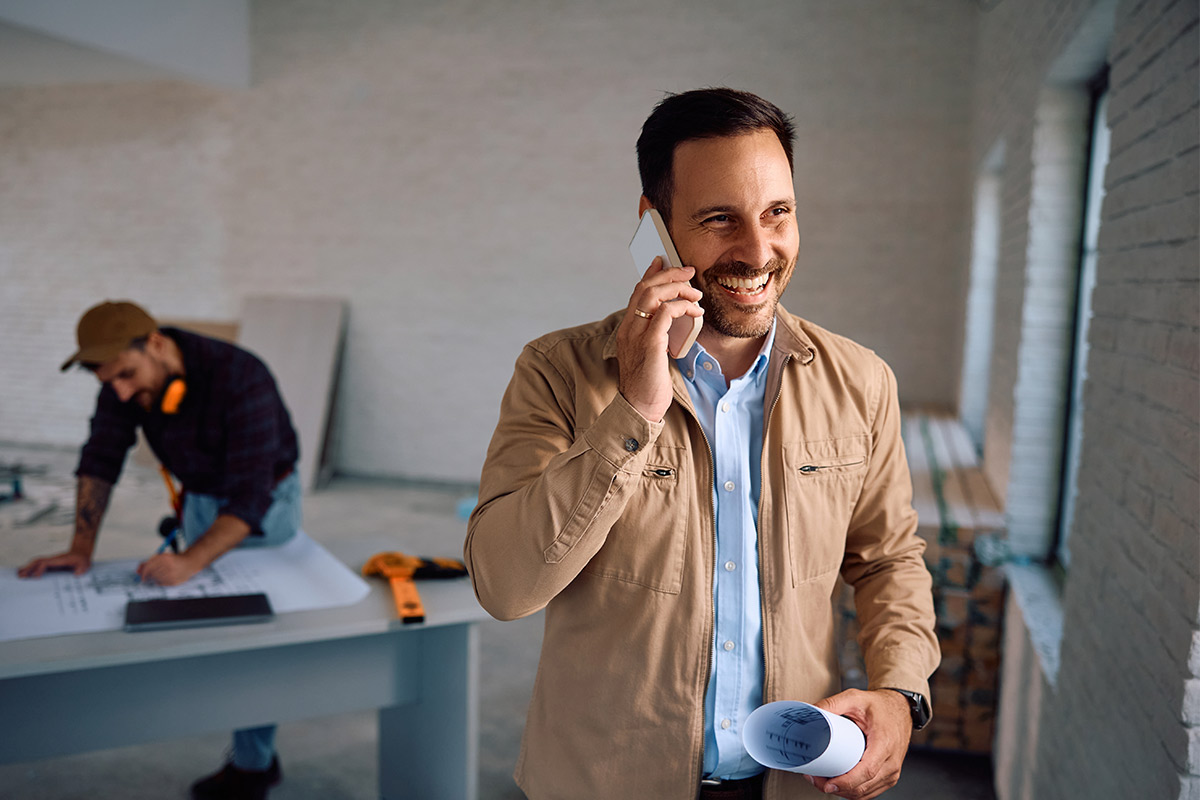 A man stands in an office, talking on a cell phone, with a desk and office equipment visible in the background.