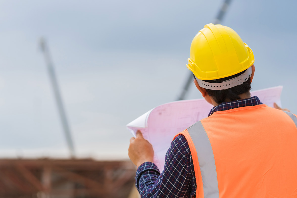 A construction worker in an orange vest and hard hat studies a blueprint, assessing the project schedule.