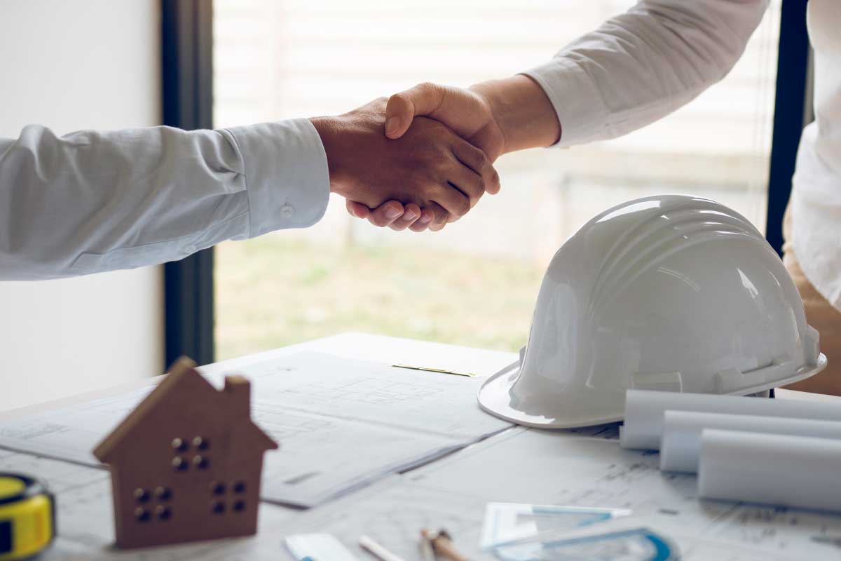 Two people shaking hands over a table with construction plans, symbolizing collaboration in project planning.