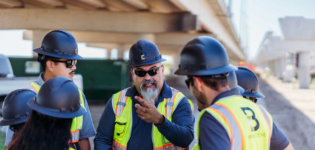 Several construction workers discussing plans on-site, equipped with safety gear and tools, surrounded by building materials.