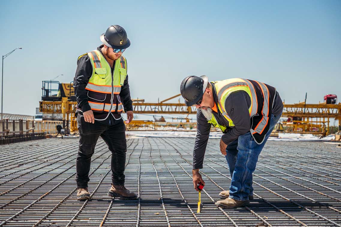 Two construction workers in hard hats standing on a steel beam at a construction site, overlooking the area below.